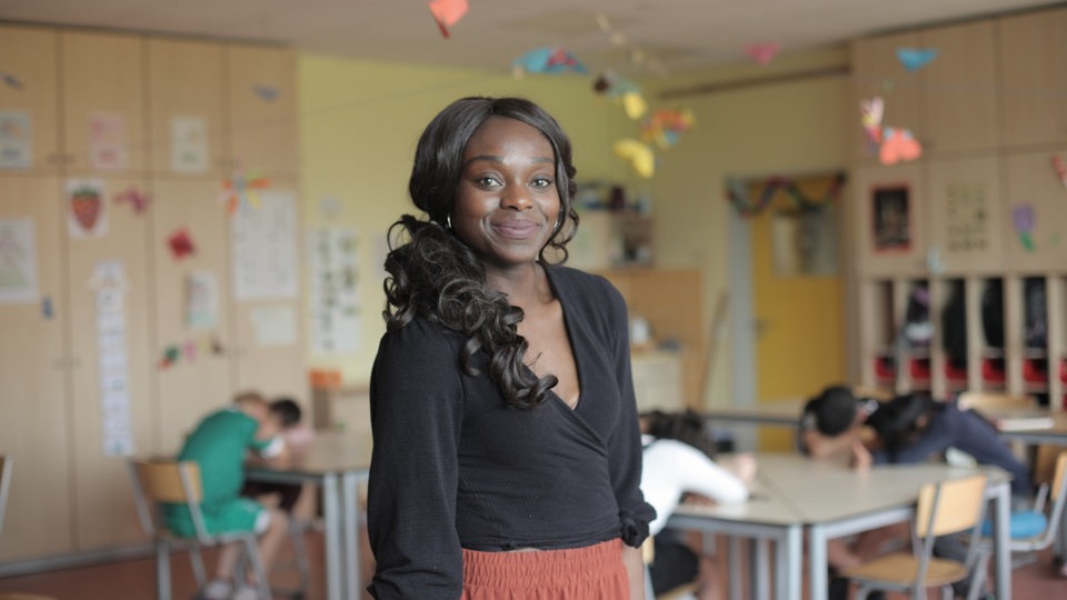 Gilda Deinhard steht in einem hellen Klassenzimmer; im Hintergrund sitzen und arbeiten Schülerinnen und Schüler.