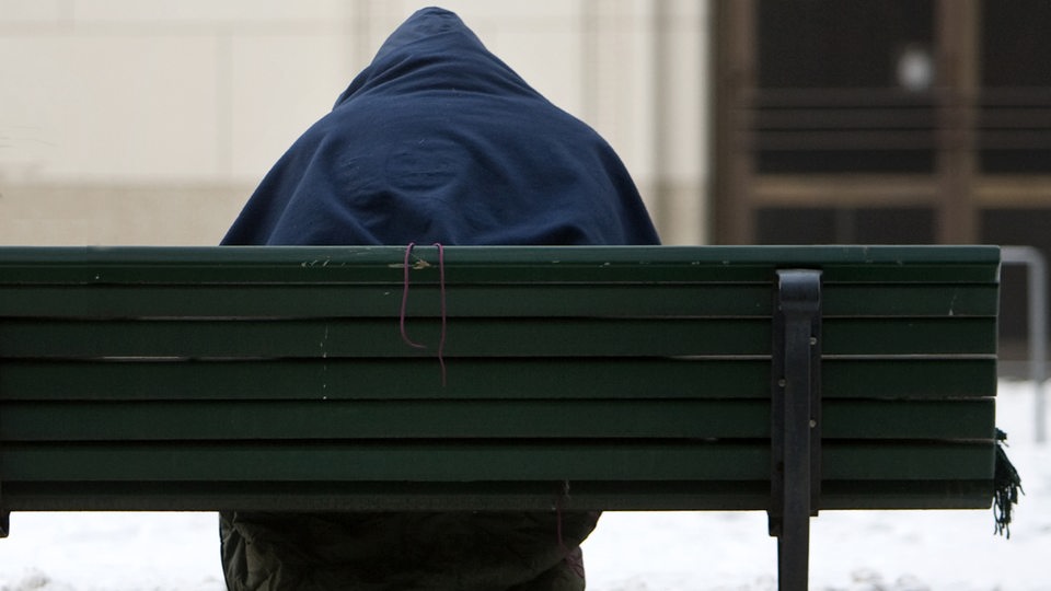 Ein Obdachloser, von hinten gesehen, sitzt im Winter mit einem Schlafsack auf einer Bank