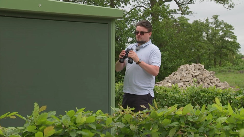 Privatdetektiv Michael Günther versteckt mit Sonnenbrille und Fernglas in der Hand, hinter eine Hecke. 