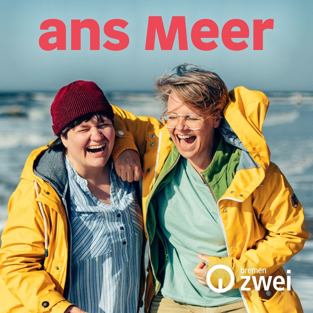 Zwei lachende Frauen mit gelben Regenmänteln, dahinter das Meer