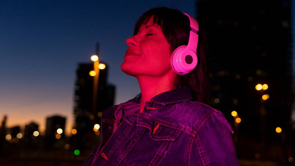 Eine Frau genießt in der Nacht mit Kopfhörern Musik vor einer städtischen Kulisse.