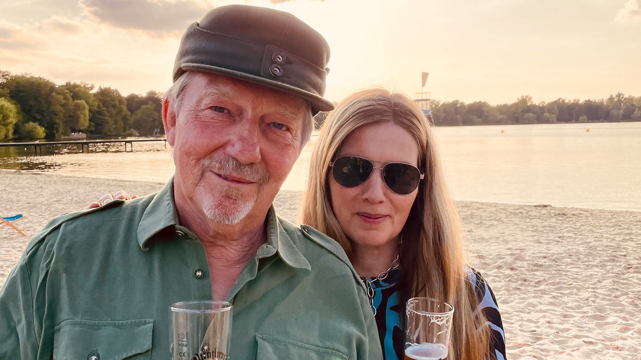 Dietmar Wischmeyer  und Tina Voß stehen mit einem Glas Bier in der Hand an einem See.